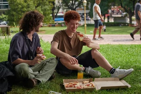 Parkta çimlerin üzerinde oturup pizza yiyen ve sohbet eden iki genç adam.