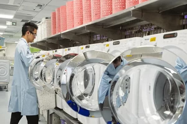 Profesyonel bir çamaşırhanede sıra halinde dizilmiş çamaşır makinelerini kontrol eden laboratuvar önlüklü bir çalışan.