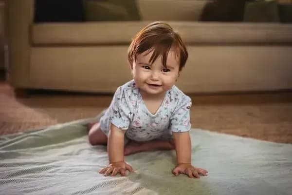 Oturma odasında bir battaniyenin üzerinde emekleyen ve kameraya gülümseyen küçük bir çocuk.